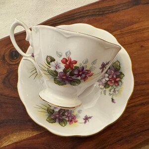 May include: A white teacup and saucer with a floral pattern featuring purple, pink, and red flowers. The flowers are surrounded by green leaves.