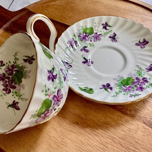 K&ouml;nnte beinhalten: Wei&szlig;es Tee-Set mit Tasse und Untertasse, verziert mit lila und gr&uuml;nen Veilchenbl&uuml;ten. Die Tasse hat einen goldfarbenen Griff. Die Untertasse ist ebenfalls goldfarben eingefasst. Ein klassisches Teeservice.