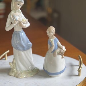 K&ouml;nnte beinhalten: Zwei Porzellanfiguren von Frauen. Eine Frau steht und h&auml;lt einen Korb mit V&ouml;geln. Die andere Frau sitzt und h&auml;lt eine Blume. Beide Figuren tragen wei&szlig;e und blaue Kleider. Die Figuren stehen auf einem Marmortablett.