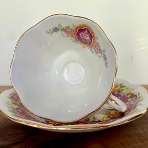 May include: White teacup and saucer set with gold trim and floral accents. The teacup is turned on its side, revealing a detailed floral pattern in pink, yellow, and blue on a burgundy background.