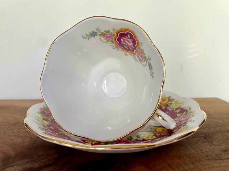 May include: White teacup and saucer set with gold trim and floral accents. The teacup is turned on its side, revealing a detailed floral pattern in pink, yellow, and blue on a burgundy background.