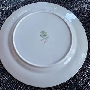 May include: A white porcelain plate with a slightly scalloped edge. The underside features a central emblem with a crown and floral design, along with text and numbers in green and red. The plate is set against a dark, textured background.