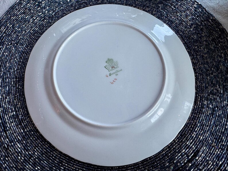 May include: A white porcelain plate with a slightly scalloped edge. The underside features a central emblem with a crown and floral design, along with text and numbers in green and red. The plate is set against a dark, textured background.
