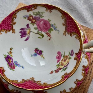 May include: A vintage teacup and saucer set with a white interior and a pink, gold, and white exterior. The rim features a pink scale pattern and gold trim. The cup and saucer are decorated with floral bouquets in shades of pink, purple, and yellow.
