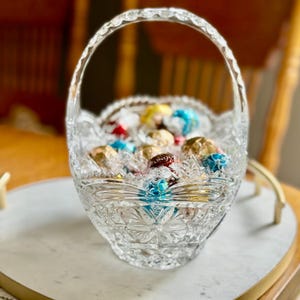 May include: A clear, cut-glass basket with a handle, filled with assorted foil-wrapped chocolates. The basket has a floral pattern on the base and a scalloped rim. The chocolates are in various colors, including gold, blue, red, and yellow.