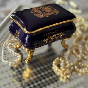 May include: A small, navy blue porcelain jewelry box with gold accents and ornate detailing. The box sits on gold-colored feet and features a decorative design on the lid. Pearl necklaces are arranged around the box on a mirrored surface.