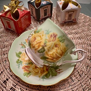 May include: A delicate teacup and saucer with a floral design featuring yellow roses on a green background. The teacup is white with a gold rim and the saucer has a scalloped edge. The teacup and saucer are sitting on a brown lace doily.