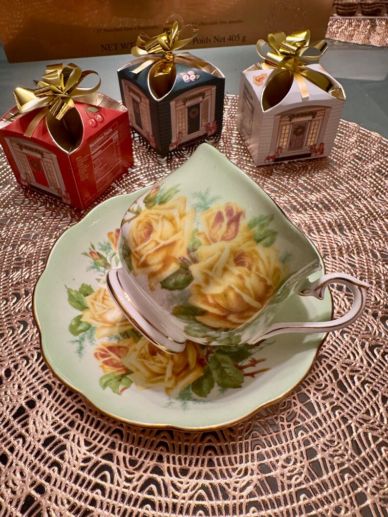 May include: A delicate teacup and saucer with a floral design featuring yellow roses on a green background. The teacup is white with a gold rim and the saucer has a scalloped edge. The teacup and saucer are sitting on a brown lace doily.