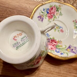 May include: A vintage teacup and saucer set with a floral pattern. The saucer has a gold rim and the teacup has a mark on the bottom that reads "Foley China Tulip Brain & Co Ltd.  English Bone China VL 705".