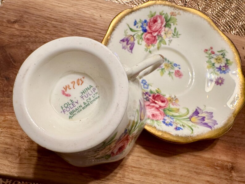 May include: A vintage teacup and saucer set with a floral pattern. The saucer has a gold rim and the teacup has a mark on the bottom that reads "Foley China Tulip Brain & Co Ltd.  English Bone China VL 705".