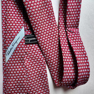 Puede incluir: Una corbata roja con un patr&oacute;n repetitivo de peque&ntilde;as figuras de elefantes en azul claro y blanco. La corbata incluye una etiqueta de tela blanca con el texto Salvatore Ferragamo y una etiqueta negra con Ferragamo Italy.