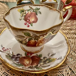 Puede incluir: Una taza de té y un platillo blancos con un borde dorado y un patrón floral con frutas y hojas. La taza y el platillo están sobre un mantel individual dorado.