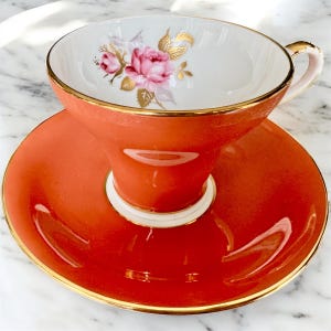 May include: A vintage teacup and saucer set with a floral design. The cup and saucer are a vibrant orange color with a gold trim. The cup has a white interior with a pink and gold floral design.