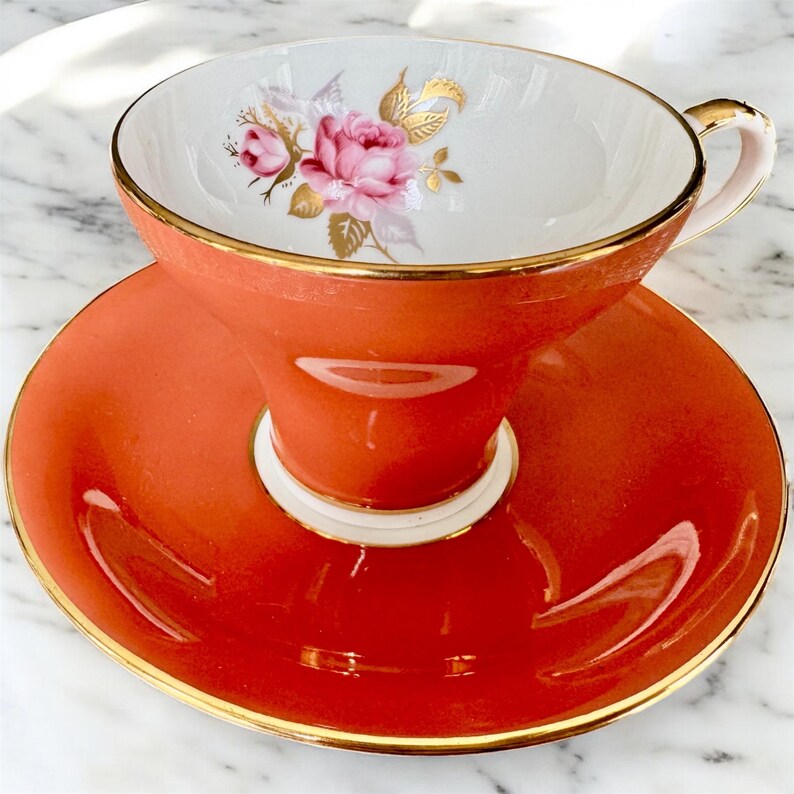 May include: A vintage teacup and saucer set with a floral design. The cup and saucer are a vibrant orange color with a gold trim. The cup has a white interior with a pink and gold floral design.