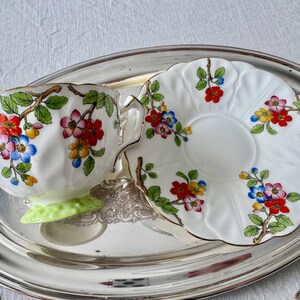 K&ouml;nnte beinhalten: Ein wei&szlig;es Tee-Set mit floralen Mustern in Rot, Blau und Gelb, akzentuiert mit goldenen Verzierungen. Die Tasse steht auf einem hellgr&uuml;nen Sockel, auf einem silbernen Tablett.