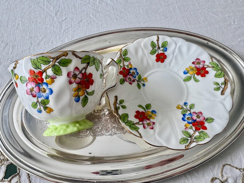 K&ouml;nnte beinhalten: Ein wei&szlig;es Tee-Set mit floralen Mustern in Rot, Blau und Gelb, akzentuiert mit goldenen Verzierungen. Die Tasse steht auf einem hellgr&uuml;nen Sockel, auf einem silbernen Tablett.