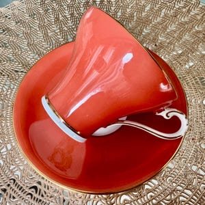 May include: A coral colored teacup and saucer with a gold trim. The cup is upside down on the saucer.