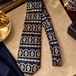 May include: A navy blue necktie featuring a repeating pattern of gold and cream-colored architectural arch motifs with orange diamond-shaped accents. The tie has a smooth, satin-like finish and a pointed tip, displaying a structured, symmetrical design.
