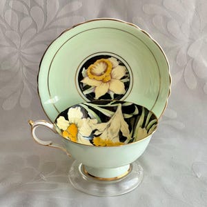 May include: A pale mint green teacup and saucer set featuring a central yellow daffodil motif against a black background. The teacup has a gold-trimmed handle and rim, with matching gold accents on the saucer edge and a scalloped saucer rim.