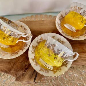 Puede incluir: Tres tazas de t&eacute; y platillos vintage. Las tazas y los platillos son blancos con detalles florales dorados y un interior amarillo brillante. Las tazas de t&eacute; est&aacute;n sobre una superficie de madera.