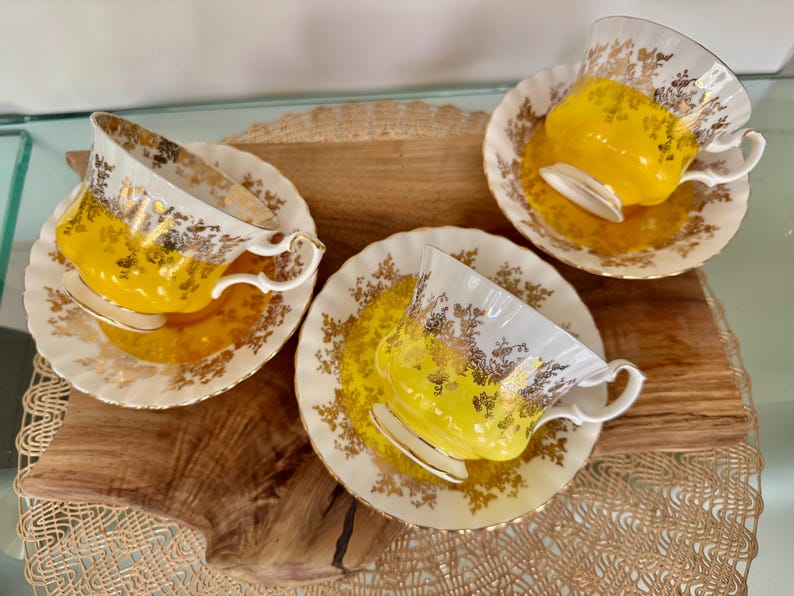 Puede incluir: Tres tazas de t&eacute; y platillos vintage. Las tazas y los platillos son blancos con detalles florales dorados y un interior amarillo brillante. Las tazas de t&eacute; est&aacute;n sobre una superficie de madera.