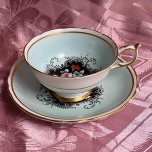 May include: A light blue porcelain teacup and saucer set featuring a gold rim, a scalloped edge, and a central black medallion with a floral design. The teacup has a curved handle and a pedestal base, with matching black floral motifs on the saucer.