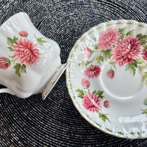 May include: A white teacup and saucer set with a floral design. The teacup has a heart-shaped handle and gold trim. The saucer and teacup are decorated with pink chrysanthemums and green leaves. The set is on a dark, textured surface.