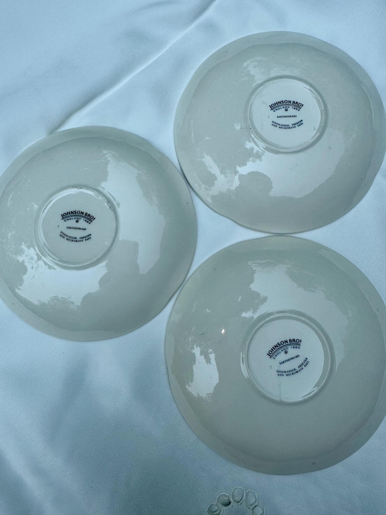 Puede incluir: Tres platillos de cer&aacute;mica blanquecina. Cada platillo tiene el texto "JOHNSON BROS ENGLAND 1883 EARTHENWARE" impreso en la base. Los platillos est&aacute;n colocados sobre una superficie blanca, resaltando su dise&ntilde;o cl&aacute;sico.