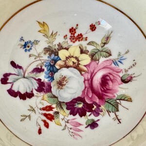 May include: A white porcelain saucer with a gold rim and a floral design. The flowers are painted in shades of pink, yellow, blue, and purple. The saucer is signed "F Howard".