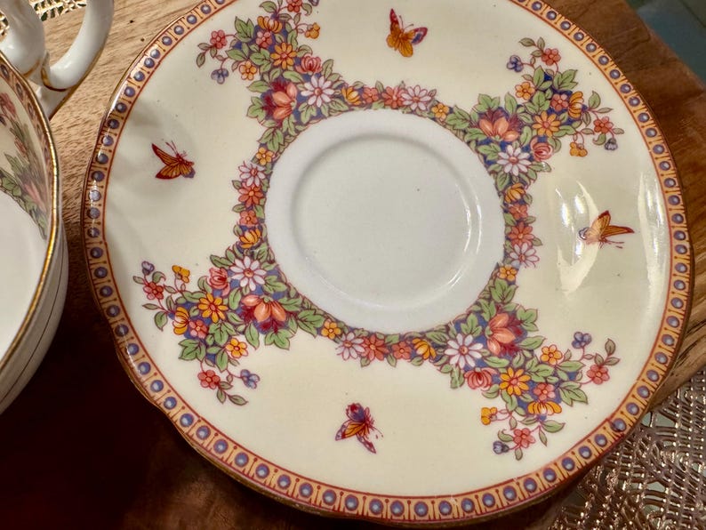 May include: A white porcelain saucer with a gold rim and a floral design featuring butterflies. The saucer has a central well for a teacup.