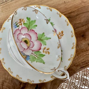 May include: A white teacup and saucer set with a floral design. The cup and saucer have a gold trim and a pink flower with green leaves.