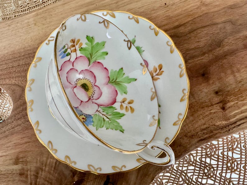 May include: A white teacup and saucer set with a floral design. The cup and saucer have a gold trim and a pink flower with green leaves.