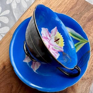 Puede incluir: Un juego de taza y platillo de t&eacute; azul y negro con un dise&ntilde;o floral. La taza y el platillo est&aacute;n decorados con una flor rosa y amarilla y hojas verdes.