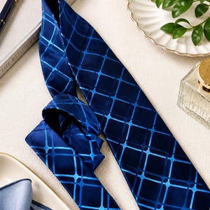 May include: A navy blue tie with a geometric grid pattern in a lighter shade of blue. The tie is made of a shiny fabric and is displayed on a light-colored surface. A matching pocket square is visible.