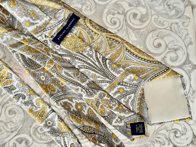 May include: A Banana Republic tie with a paisley pattern in shades of yellow, gray, and white. The tie is made in the USA and features the brand's label.
