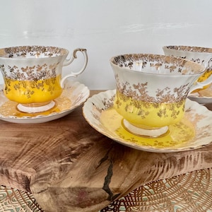 Puede incluir: Tres tazas de t&eacute; y platillos vintage. Las tazas tienen un dise&ntilde;o floral blanco y dorado con una base amarilla. Los platillos son blancos con un estampado floral dorado. Las tazas de t&eacute; est&aacute;n sobre una superficie de madera.