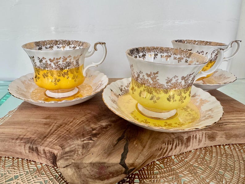 Puede incluir: Tres tazas de t&eacute; y platillos vintage. Las tazas tienen un dise&ntilde;o floral blanco y dorado con una base amarilla. Los platillos son blancos con un estampado floral dorado. Las tazas de t&eacute; est&aacute;n sobre una superficie de madera.