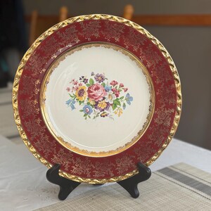 May include: A vintage decorative plate with a red border featuring a gold floral pattern. The center of the plate is white with a floral bouquet design in pink, yellow, blue, and purple. The plate is displayed on a black stand.