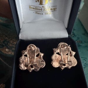 Op de afbeelding: Een paar goudkleurige clip-on oorbellen met een sierlijk bloemmotief, gepresenteerd in een donkerblauwe fluwelen doos. De doos toont het logo van Bradford Exchange.
