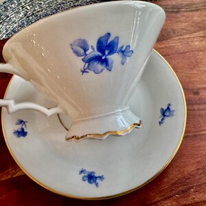 May include: A white teacup and saucer with a blue floral design. The cup has a scalloped edge and the saucer has a gold rim.