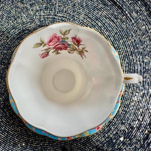 K&ouml;nnte beinhalten: Eine wei&szlig;e Teetasse mit blauem und goldenem Rand. Die Teetasse zeigt ein Blumenmuster mit rosa Rosen und gr&uuml;nen Bl&auml;ttern.