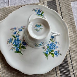 May include: A white teacup and saucer set with a floral design. The teacup has the words "Forget-Me-Not Royal Albert Bone China England" printed on the bottom.