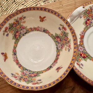 May include: A set of two vintage teacups and saucers with a floral and butterfly design. The cups and saucers are white with a gold rim and feature a delicate floral pattern in shades of pink, blue, and yellow. The design also includes small butterflies in shades of orange and brown.