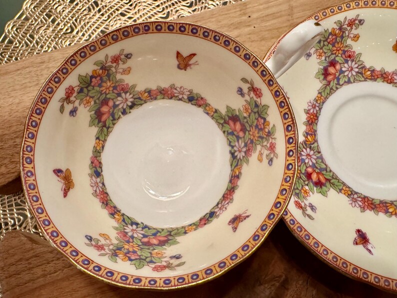 May include: A set of two vintage teacups and saucers with a floral and butterfly design. The cups and saucers are white with a gold rim and feature a delicate floral pattern in shades of pink, blue, and yellow. The design also includes small butterflies in shades of orange and brown.