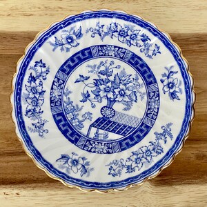 May include: A white and blue decorative plate with a floral design. The plate features a blue floral pattern around the rim and a central design of flowers in a vase. The plate has a scalloped edge with a gold trim.