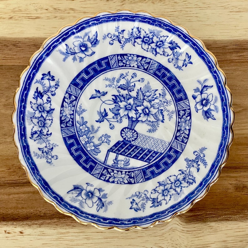 May include: A white and blue decorative plate with a floral design. The plate features a blue floral pattern around the rim and a central design of flowers in a vase. The plate has a scalloped edge with a gold trim.