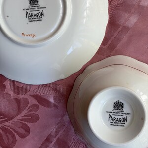 May include: Two white Paragon fine bone china pieces, a saucer and a bowl, each featuring a black backstamp that reads By Appointment To Her Majesty The Queen China Potters Paragon Fine Bone China England Regd. The saucer has a handwritten mark.