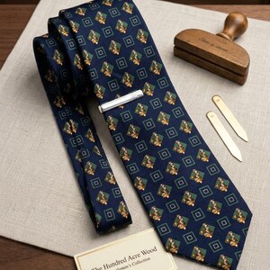 K&ouml;nnte beinhalten: Eine marineblaue Krawatte mit einem sich wiederholenden Muster von Figuren in gr&uuml;nen und goldenen Rauten. Eine silberne Krawattenklammer ist angebracht. The Hundred Acre Wood - A Gentleman's Collection ist auf einer kleinen Karte aufgedruckt.
