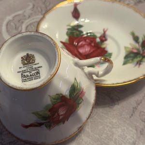 May include: White Paragon fine bone china teacup and saucer set. The teacup and saucer feature a red rose floral design with green leaves and gold trim. The teacup has a gold handle and the saucer has a scalloped edge.