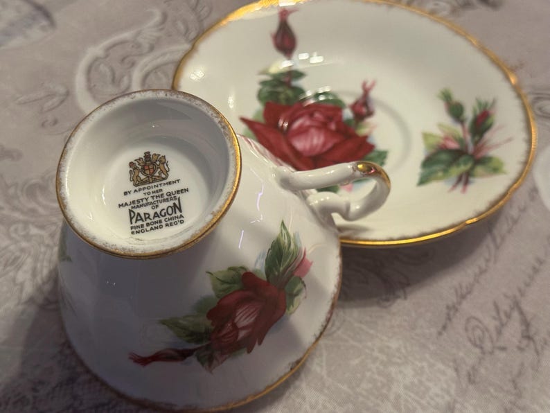 May include: White Paragon fine bone china teacup and saucer set. The teacup and saucer feature a red rose floral design with green leaves and gold trim. The teacup has a gold handle and the saucer has a scalloped edge.